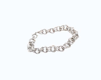 May include: A silver-colored bracelet with a double-strand chain design. The bracelet features a series of interconnected, rounded links, creating a textured and eye-catching accessory. The clasp is visible, securing the bracelet.