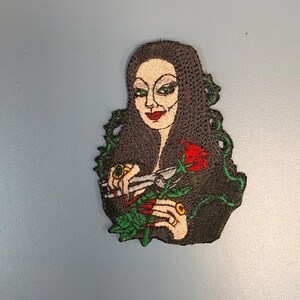 Könnte beinhalten: Gestickter Aufnäher mit einer Frau mit langen schwarzen Haaren, die ein schwarzes Kleid trägt und eine rote Rose hält. Sie hat einen finsteren Ausdruck und trägt goldene Ringe an den Fingern.