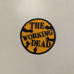 May include: Round patch with a yellow background and black border. The text "THE WORKING DEAD" is in black, with black hands reaching out from the edges. The patch is likely for clothing or accessories.