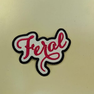 May include: Embroidered patch with the word "Feral" in red script lettering. The text is on a white background, outlined with a black border and a white border. The patch is likely for clothing or accessories.