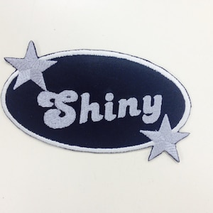 May include: A navy blue oval patch with white stitching and the word "Shiny" in a white, retro font. Two white stars are on either side of the patch.