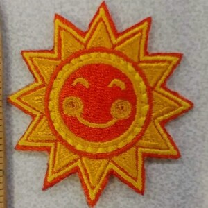 May include: Embroidered patch featuring a smiling sun with a cheerful expression. The sun has a bright orange face and yellow rays, creating a vibrant and playful design. This patch is suitable for clothing, bags, or other fabric items.