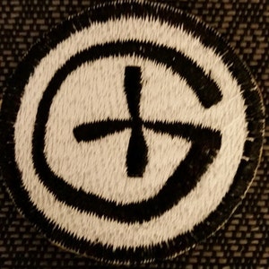 May include: Black and white embroidered patch with a circle and a cross inside. The circle is outlined in black and the cross is black.