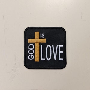 Könnte beinhalten: Ein schwarzer Stoff-Patch mit einem goldenen gestickten Kreuz und dem Text "GOD IS LOVE" in weißer Stickerei.