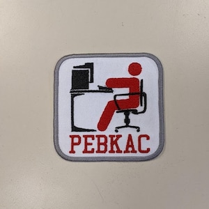May include: A gray and white embroidered patch with a red stick figure sitting at a desk with a computer. The text "PEBKAC" is written in red below the figure.