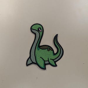May include: A green cartoon dinosaur patch with a long neck and tail. The patch has a black outline and a gray border. The dinosaur has a few brown spots on its back and is set against a white background.