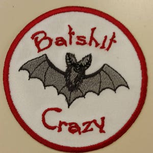 May include: A white embroidered patch with a red border. The patch features a black bat with gray wings and the text "Bats**h**it Crazy" in red.