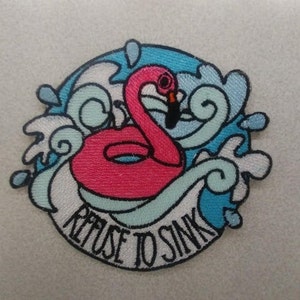 Könnte beinhalten: Gestickter Aufnäher mit einem rosa Flamingo-Schwimmring in blauem Wasser mit weißen Wellen. Der Text "Refuse to Sink" ist auf einem weißen Banner geschrieben.