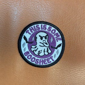 May include: Round embroidered patch with the text "THIS IS SOME BOOSHEET" in purple and white. The patch features a cartoon ghost with an angry expression and two small black knives. The patch has a black border and is set against a brown background.