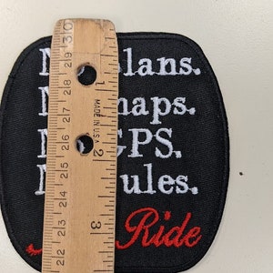 May include: Black and white embroidered patch with the text "No plans. No maps. PS. Rules. Ride" in white lettering. The word "Ride" is in red lettering. The patch is approximately 3 inches in diameter.
