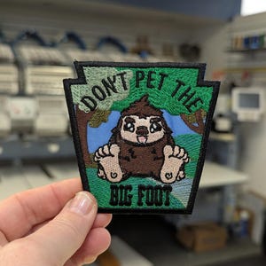 May include: Embroidered patch with a green and black border, featuring a cartoon Bigfoot with the text "DON'T PET THE BIG FOOT." The Bigfoot is brown with large feet and a smiling face, set against a blue and green background.