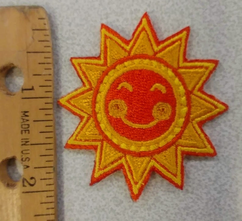 May include: Embroidered patch featuring a smiling sun design. The sun has a bright orange center with a yellow outline and rays. The patch is approximately 2 inches in diameter. The patch is made in the U.S.A.