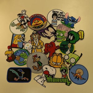 May include: A collection of colorful embroidered patches featuring cartoon characters. The patches include characters from Looney Tunes, The Simpsons, and other animated shows. The patches are round and rectangular, with various colors and designs.