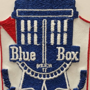 May include: A blue and white embroidered patch featuring a blue box with the text "Blue Box" and a blue ribbon with the text "Beer It" in white. The patch has a red and white diagonal stripe in the background.