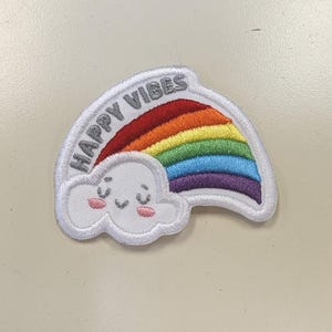 May include: Embroidered patch featuring a rainbow and a smiling cloud. The rainbow displays vibrant colors, and the text "HAPPY VIBES" is arched above it. The patch has a white border and is designed for clothing or accessories.