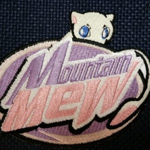May include: A purple and pink embroidered patch with the words "Mountain Dew" in a white outline. A white cartoon cat with blue eyes is peeking out from behind the text.