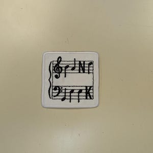 May include: Square white patch with black musical notes and symbols. The patch features a treble clef, bass clef, and musical notes. The letters "N" and "K" are also visible. The patch is centered on a light beige surface.