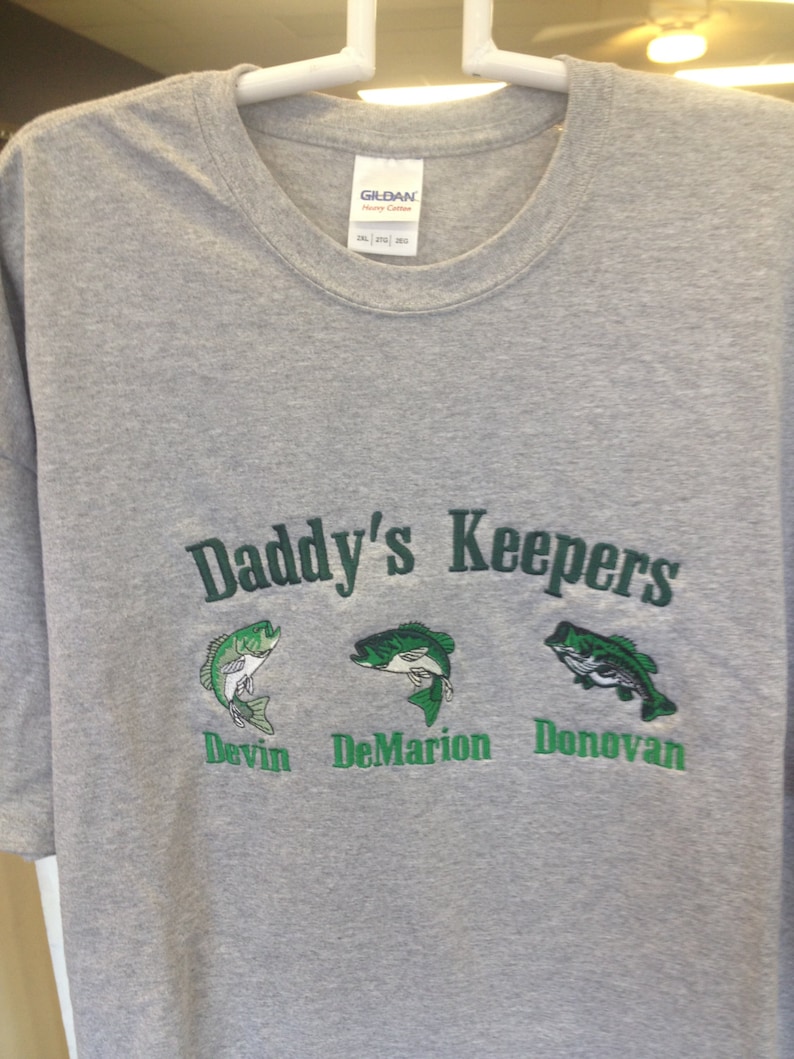 May include: Gray t-shirt with the text "Daddy's Keepers" in green. Three green fish are embroidered below the text, each with a name underneath: Devin, DeMarion, and Donovan.