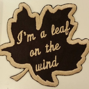 May include: A brown felt leaf-shaped patch with beige stitching. The text "I'm a leaf on the wind" is embroidered in beige thread.