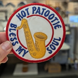 May include: Embroidered patch with a white background, a red border, and blue text that reads "HOT PATOOTIE BLESS MY SOUL." A yellow saxophone is in the center. The patch is round.