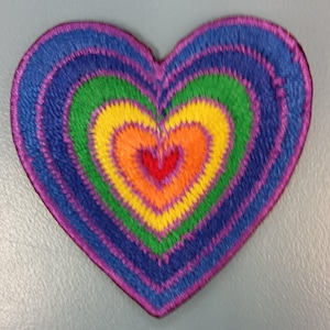 May include: A rainbow heart-shaped patch with a smaller red heart in the center. The patch is made of embroidered fabric and has a purple, blue, green, yellow, orange, and red color scheme.