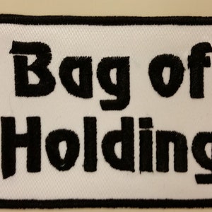 Peut inclure: Écusson brodé blanc avec des points noirs. L'écusson porte l'inscription "Bag of Holding" en caractères gras et carrés.