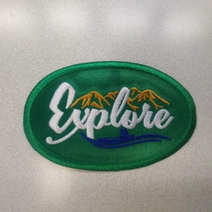 May include: Green oval embroidered patch with white text that says "Explore" and a gold mountain range behind the text. A blue boat is below the text and a blue wave is below the boat.