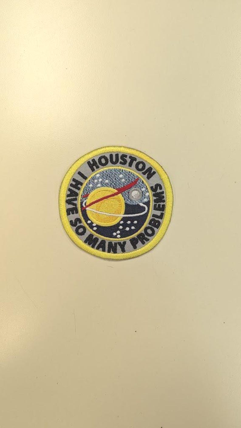 Puede incluir: Parche bordado circular con la frase "Houston, tengo muchos problemas". El parche presenta un sol amarillo, un planeta y una l&iacute;nea roja. Tiene un borde amarillo y un fondo azul marino.