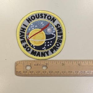 Puede incluir: Parche bordado con el texto "HOUSTON I HAVE SO MANY PROBLEMS". El parche presenta un borde amarillo y un dise&ntilde;o de un planeta, estrellas y una l&iacute;nea roja. Una regla de madera mide hasta 10 cm.