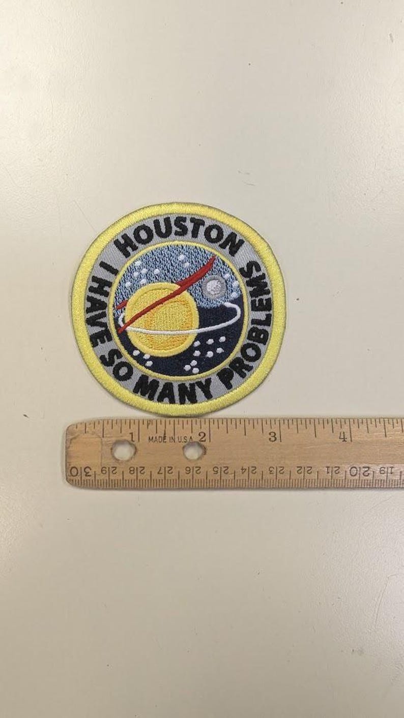 Puede incluir: Parche bordado con el texto "HOUSTON I HAVE SO MANY PROBLEMS". El parche presenta un borde amarillo y un dise&ntilde;o de un planeta, estrellas y una l&iacute;nea roja. Una regla de madera mide hasta 10 cm.