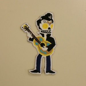 May include: An embroidered patch of a skeleton playing a yellow guitar. The skeleton is wearing a black jacket and blue jeans. The patch is white with black outlines.