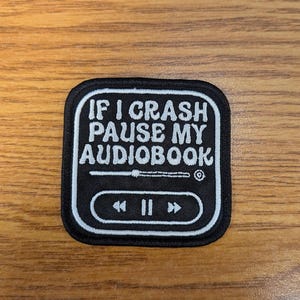 May include: A black square patch with white text that reads "IF I CRASH PAUSE MY AUDIOBOOK." The patch has a white border and features a graphic of a media player with rewind, play, and fast forward buttons. The patch is on a wooden surface.