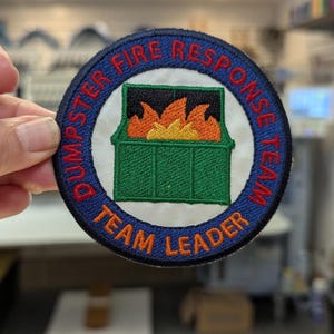 May include: Embroidered patch with a blue border and the words "DUMPSTER FIRE RESPONSE TEAM LEADER" in red and white. The patch features a green dumpster with orange and yellow flames inside.