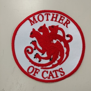 Peut inclure: Un écusson circulaire blanc avec une bordure rouge brodée. Au centre, un chat à trois têtes rouge avec des contours noirs, entouré du texte "MOTHER OF CATS".