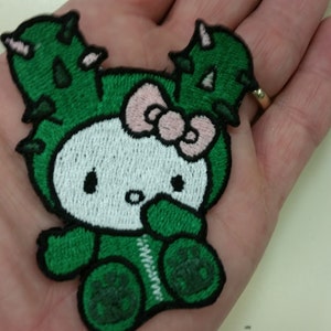 May include: Green embroidered patch of a cartoon character wearing a cactus costume with a pink bow.