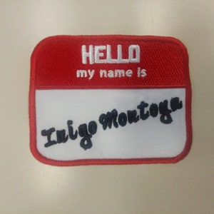 May include: Red and white name tag with the text "HELLO my name is" and the name "Inigo Montoya" written in black cursive.