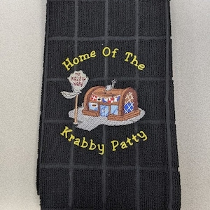 May include: Black kitchen towel with yellow embroidered text that reads "Home of the Krusty Krab" and "Krabby Patty". The embroidery also includes a cartoon image of the Krusty Krab restaurant.