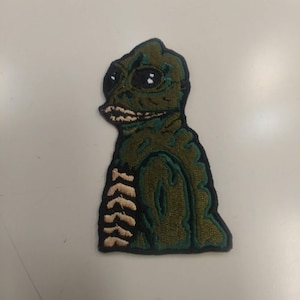 May include: Green embroidered patch of a creature with large eyes and a toothy grin. The creature has a scaly texture and is wearing a jacket with a zipper.