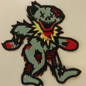 May include: A green, red, and yellow embroidered patch of a cartoon bear with a skull face and blood dripping from its mouth. The bear is walking with its arms outstretched.