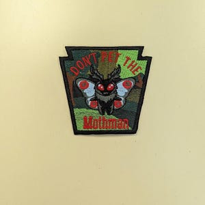 May include: Embroidered patch featuring a moth-like creature with red eyes and blue and red wing accents. The patch is in a shield shape with the text "DON'T PET THE Mothman" in red and green.