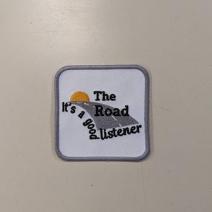 May include: White embroidered patch with a gray road and a yellow sun. The text reads "The Road It's a good listener."