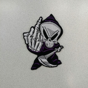 May include: An embroidered patch featuring a skeleton wearing a purple hooded robe and holding a scythe. The skeleton is giving the middle finger.