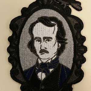 May include: Embroidered patch featuring a portrait of Edgar Allan Poe in a black ornate frame, with a black raven perched above. The portrait is in shades of gray and black, with a blue accent on the clothing.