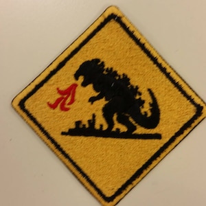 May include: Yellow diamond-shaped warning sign with a black border. The sign depicts a black silhouette of a Godzilla-like monster with red flames coming from its mouth.