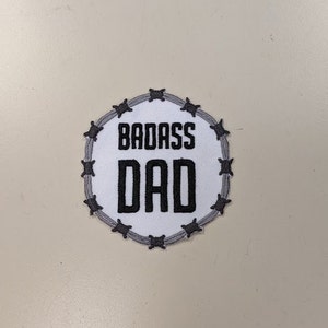May include: White round patch with a barbed wire border. The patch says "BADASS DAD" in black text.