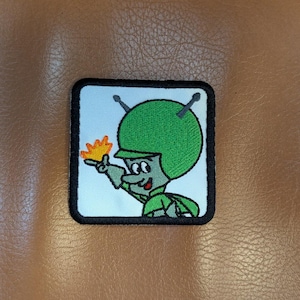 May include: Embroidered patch featuring a cartoon alien character in a green helmet and uniform, holding a yellow flame. The patch has a black border and is set against a light blue background.