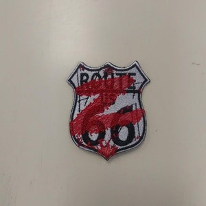 May include: A white and black embroidered patch shaped like a shield with the text "ROUTE 66" in black. The patch is covered in red paint splatters.