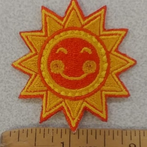 May include: Embroidered patch featuring a smiling sun with a cheerful expression. The sun has a bright orange center and yellow rays. The patch is approximately 2 inches in diameter. The text "MADE IN U.S.A." is visible.