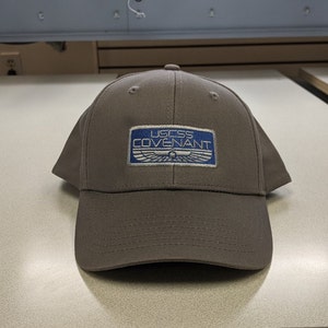 May include: A brown baseball cap with a blue and white embroidered patch that reads "UGCSS COVENANT".