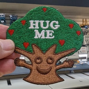 May include: Embroidered patch featuring a smiling tree with outstretched arms, a green leafy top, and the words "HUG ME" in white. Red heart accents are scattered throughout the design. The patch is held up by a hand.